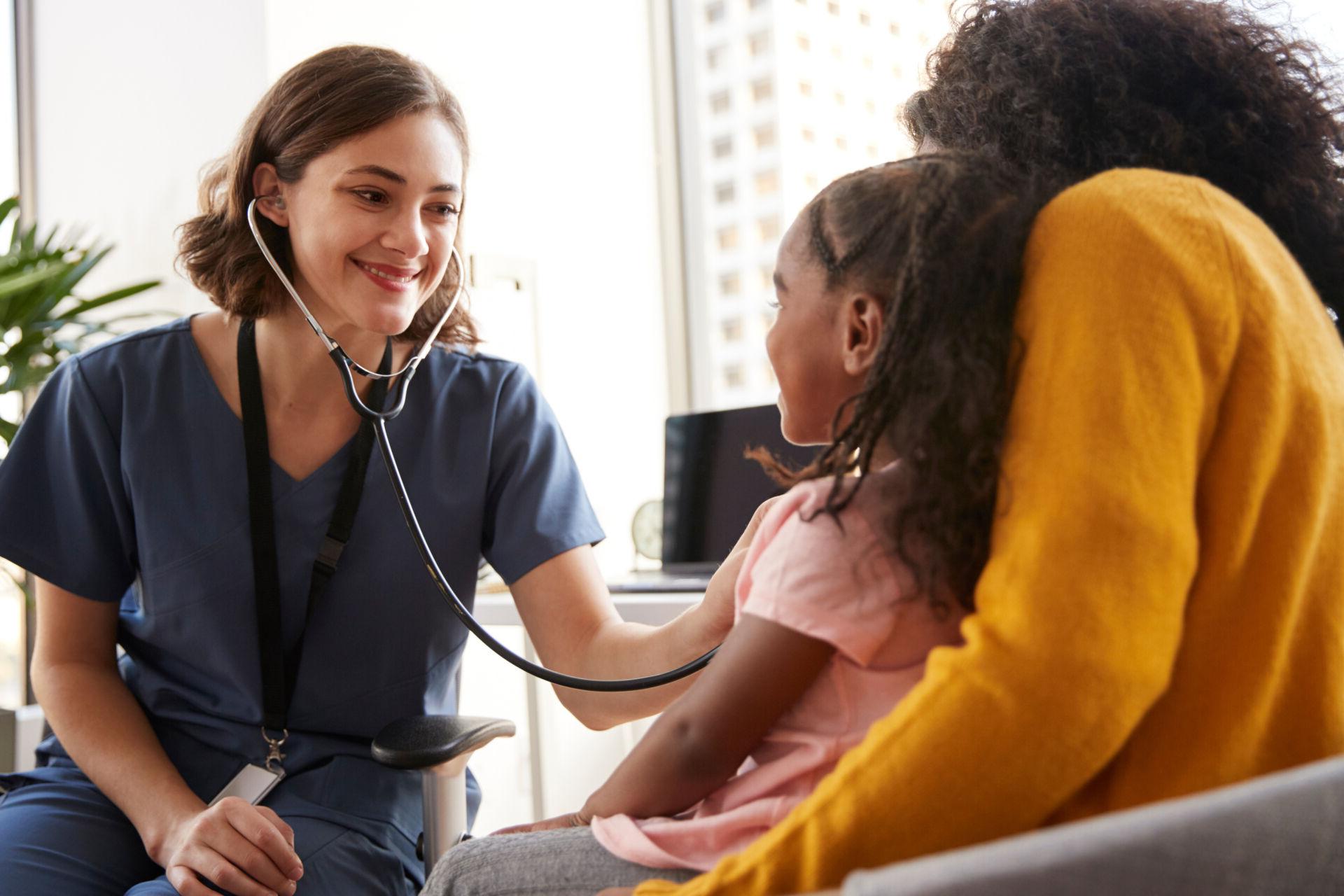 Female Pediatrician Wearing Scrubs Listening To Girls Chest With Stethoscope In Hospital Office